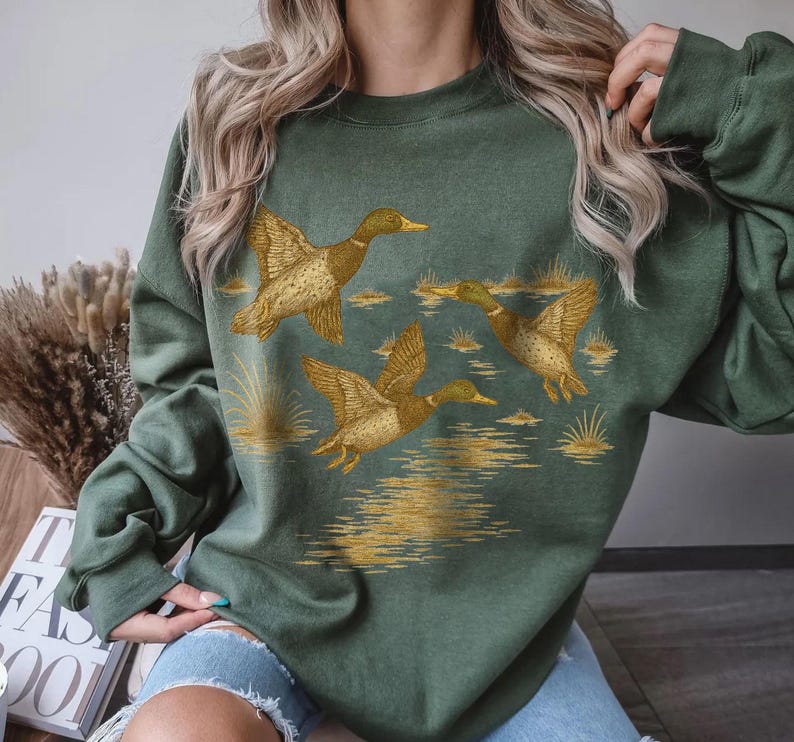 May include: Green sweatshirt featuring a detailed illustration of three ducks in flight over a body of water. The ducks are depicted in shades of brown and gold, with the water and reeds also in gold. The sweatshirt is a solid green color.