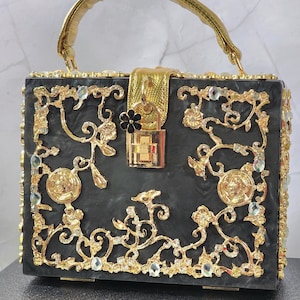 May include: A black box handbag with gold detailing and a gold handle. The bag is decorated with gold filigree, sparkling rhinestones, and a gold clasp. A small black flower charm hangs from the clasp.