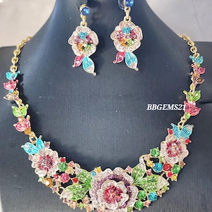 Pode incluir: Um conjunto de colar e brincos dourados com desenhos florais. O colar e os brincos são adornados com pedras preciosas coloridas e folhas verdes. Os brincos têm topos de pedras preciosas azuis. O texto "BBGEMS21" é visível.