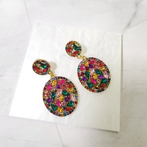May include: A pair of gold tone dangle earrings with oval shaped settings. The settings are covered in colorful rhinestones in shades of pink, green, blue, red, and yellow.