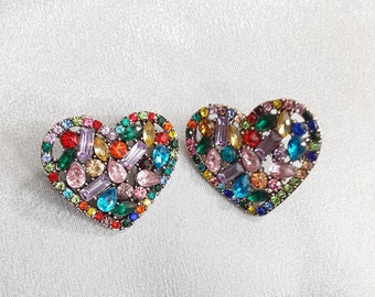 Op de afbeelding: Een paar hartvormige oorbellen met een zilverkleurige metalen setting en een kleurrijk assortiment aan strass steentjes. De strass steentjes zijn in verschillende vormen en maten, waaronder rond, ovaal en rechthoekig.