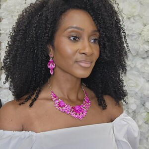 May include: A woman wearing a bright pink necklace and matching earrings. The jewellery features a floral design with gold detailing. The woman is wearing a white off-the-shoulder top, and her dark curly hair frames her face. The background is a wall of white flowers.