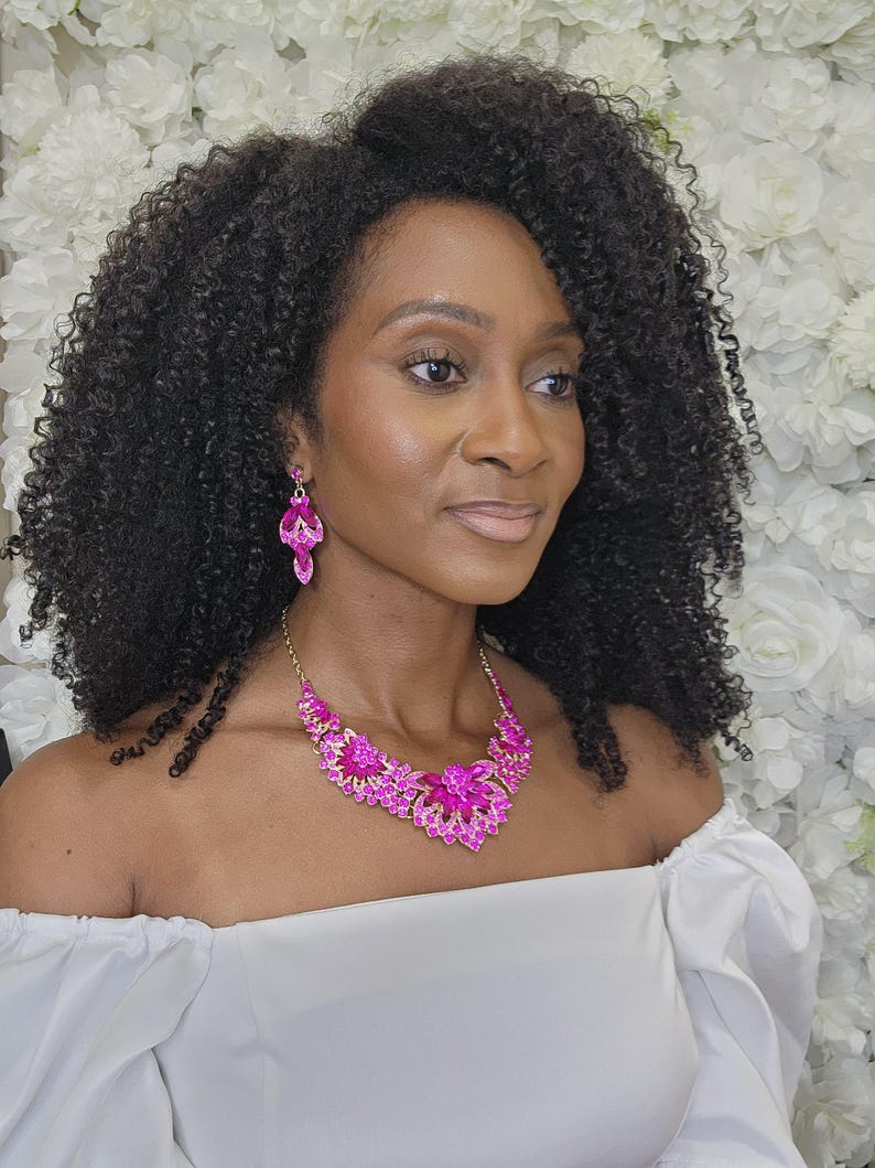 May include: A woman wearing a bright pink necklace and matching earrings. The jewellery features a floral design with gold detailing. The woman is wearing a white off-the-shoulder top, and her dark curly hair frames her face. The background is a wall of white flowers.