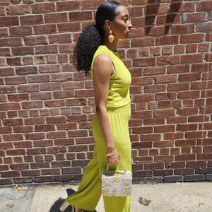 May include: A woman wearing a bright green, sleeveless, ribbed crop top and matching wide-leg pants. She is carrying a silver and gold box clutch.