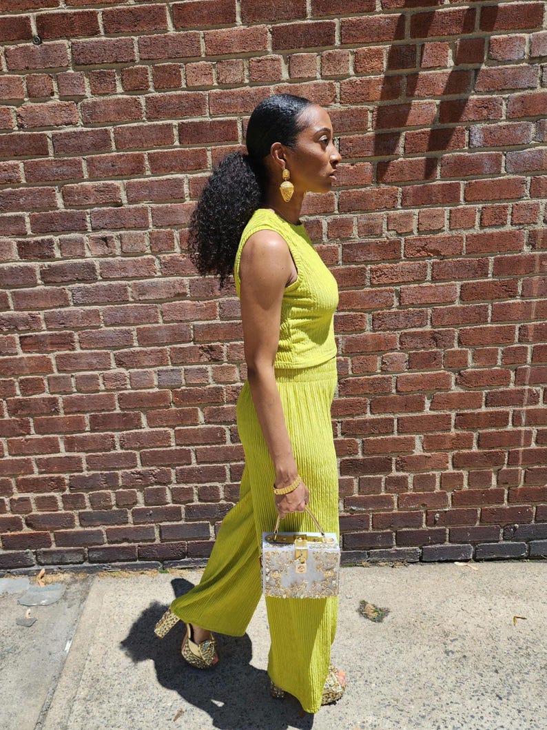 May include: A woman wearing a bright green, sleeveless, ribbed crop top and matching wide-leg pants. She is carrying a silver and gold box clutch.