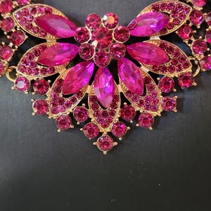 May include: A gold-coloured necklace featuring a central floral design. The necklace is embellished with numerous fuchsia-coloured rhinestones in various shapes and sizes, creating a sparkling and ornate appearance. The background is a solid black.