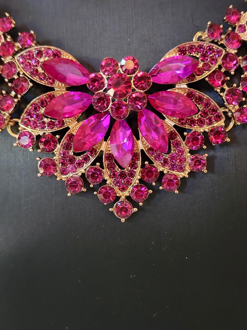 May include: A gold-coloured necklace featuring a central floral design. The necklace is embellished with numerous fuchsia-coloured rhinestones in various shapes and sizes, creating a sparkling and ornate appearance. The background is a solid black.
