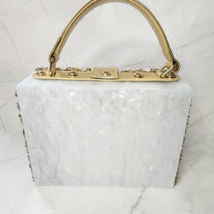 May include: A white rectangular box clutch with a gold top handle and gold embellishments. The box is made of a white marbled material.