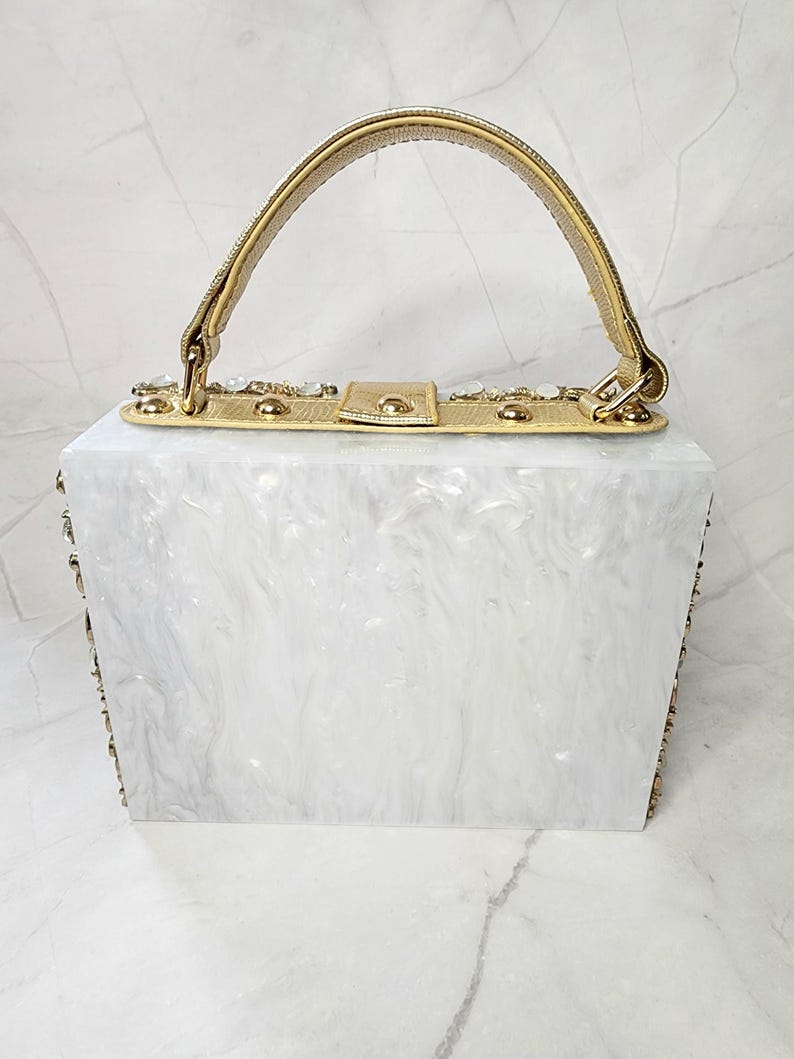 May include: A white rectangular box clutch with a gold top handle and gold embellishments. The box is made of a white marbled material.