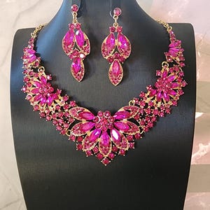 May include: A gold-coloured necklace and earring set adorned with fuchsia gemstones. The necklace has a floral design with a large central flower and smaller details. The earrings are teardrop-shaped, matching the necklace. The jewellery is displayed on a black mannequin.