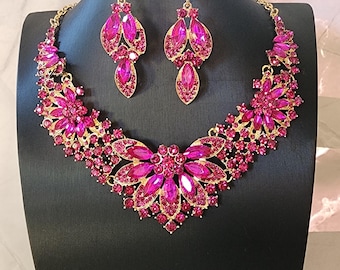 May include: A gold-toned necklace and earring set with fuchsia-colored gemstones. The necklace features a floral design with a large central flower and smaller accents. The earrings are teardrop-shaped, complementing the necklace's design. The jewelry is displayed on a black mannequin.