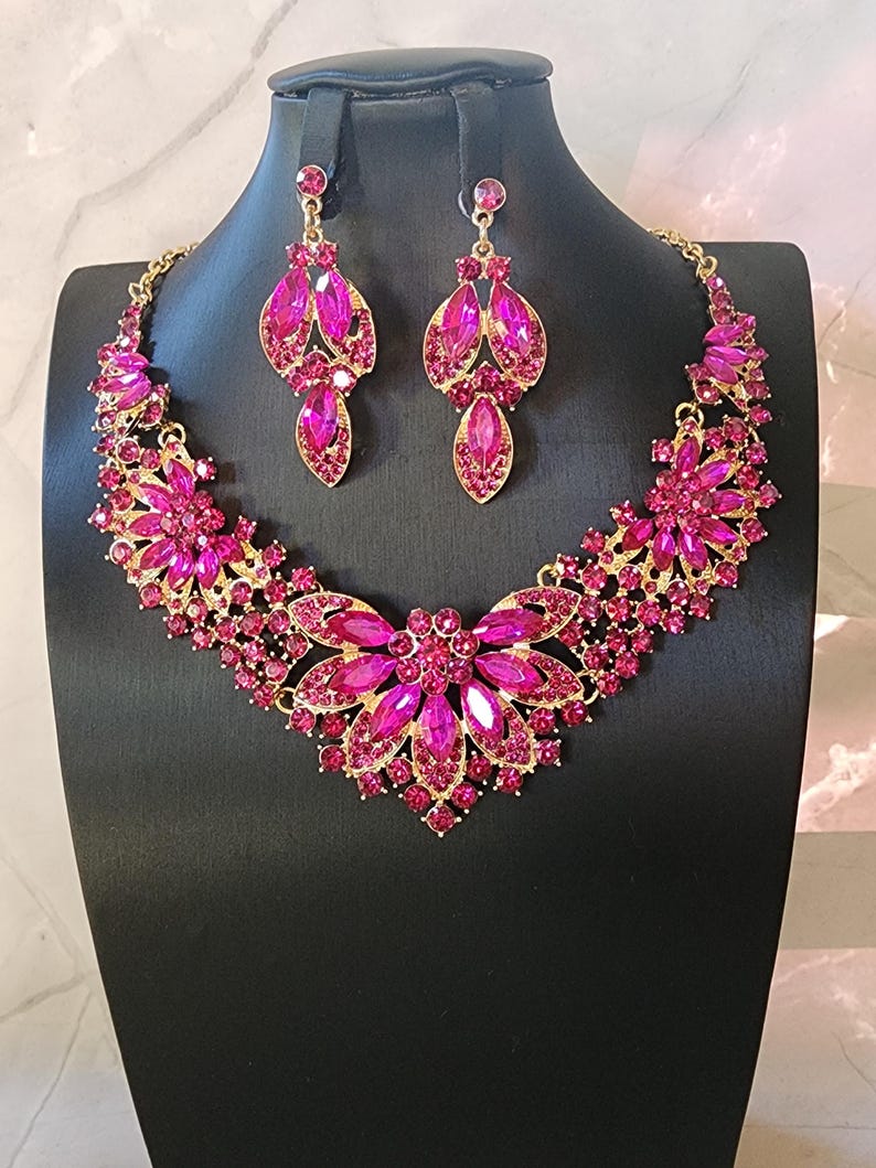 May include: A gold-coloured necklace and earring set adorned with fuchsia gemstones. The necklace has a floral design with a large central flower and smaller details. The earrings are teardrop-shaped, matching the necklace. The jewellery is displayed on a black mannequin.