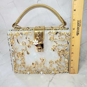May include: A white clutch purse with a gold handle and intricate gold floral design embellished with rhinestones. The purse has a gold lock and keyhole.