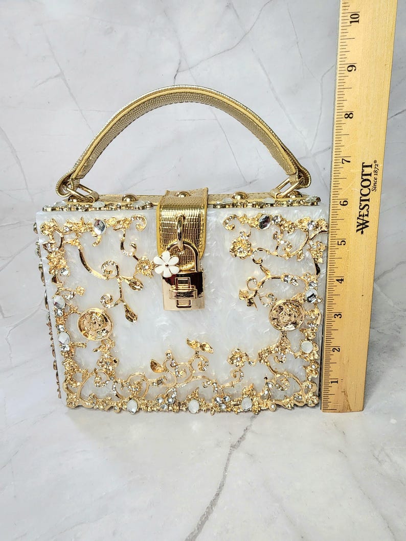 May include: A white clutch purse with a gold handle and intricate gold floral design embellished with rhinestones. The purse has a gold lock and keyhole.