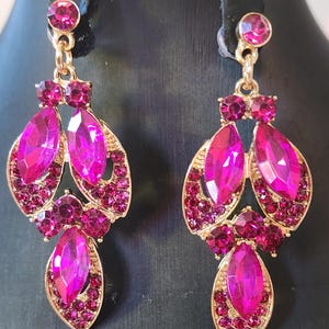 May include: A pair of gold-coloured earrings with a cascading design. Each earring features large, fuchsia-coloured, teardrop-shaped gems and smaller, round, fuchsia-coloured gems. The earrings are set against a black background.