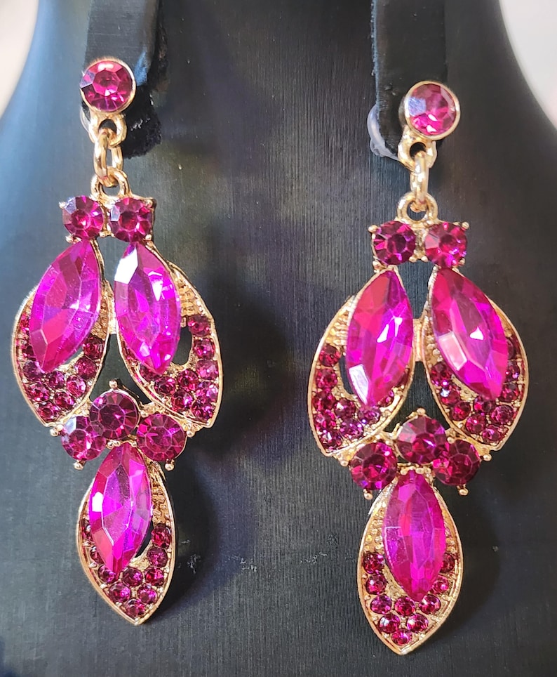 May include: A pair of gold-coloured earrings with a cascading design. Each earring features large, fuchsia-coloured, teardrop-shaped gems and smaller, round, fuchsia-coloured gems. The earrings are set against a black background.