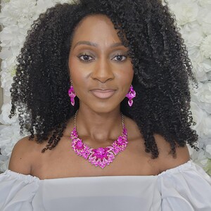 May include: A woman wearing a vibrant pink crystal necklace and matching earrings. The necklace has a floral design with a central flower and surrounding petals. The earrings are a dangling design. The woman is wearing a white off-the-shoulder top.