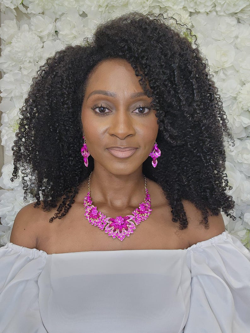 May include: A woman wearing a vibrant pink crystal necklace and matching earrings. The necklace has a floral design with a central flower and surrounding petals. The earrings are a dangling design. The woman is wearing a white off-the-shoulder top.