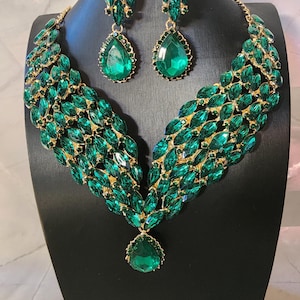 Puede incluir: Un deslumbrante conjunto de joyas verde esmeralda, que incluye un collar y pendientes a juego. El collar presenta un diseño en cascada de piedras preciosas verdes con un gran colgante en forma de lágrima. Los pendientes tienen un diseño similar. Exhibido en un maniquí negro.