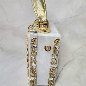 May include: A white box clutch with a gold handle and gold embellishments. The box is decorated with clear rhinestones.