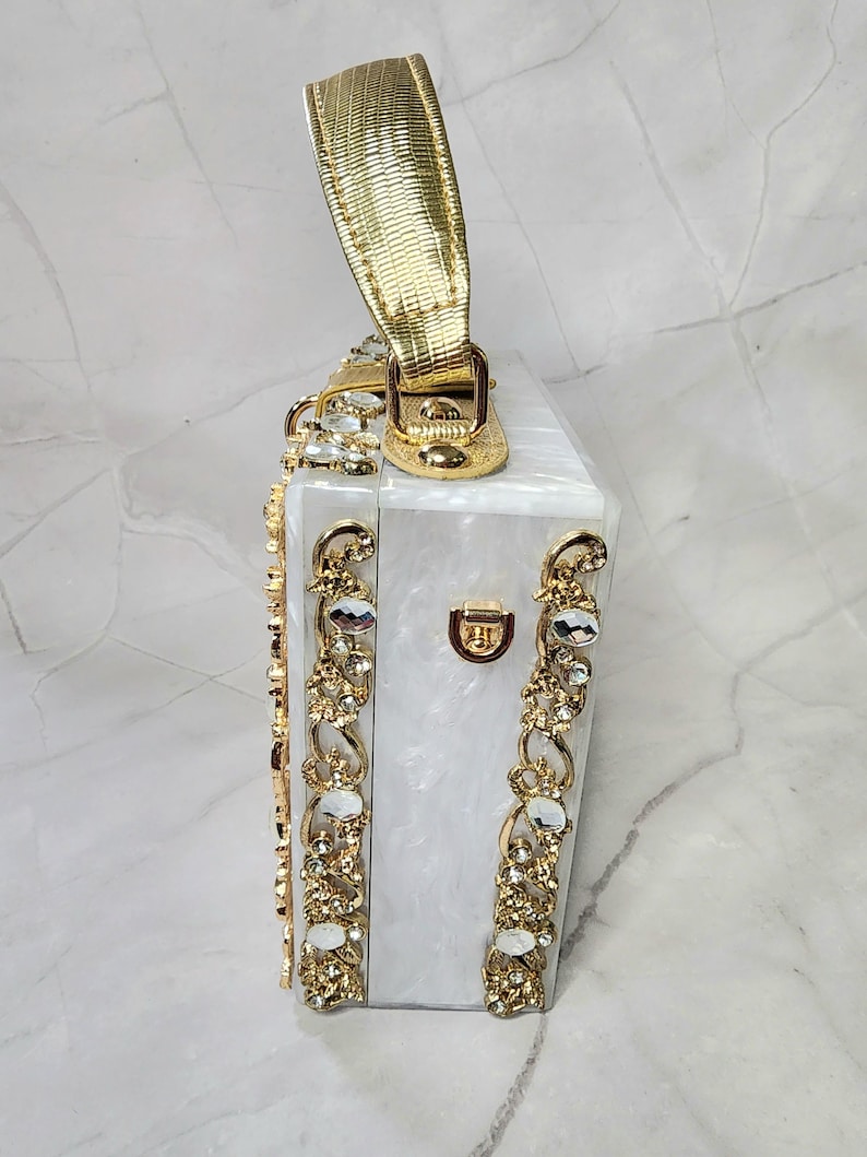 May include: A white box clutch with a gold handle and gold embellishments. The box is decorated with clear rhinestones.