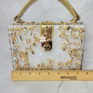 May include: A white box clutch with a gold handle and ornate gold and crystal embellishments. The clutch has a gold lock and a small white flower charm. The clutch is 6 inches wide.