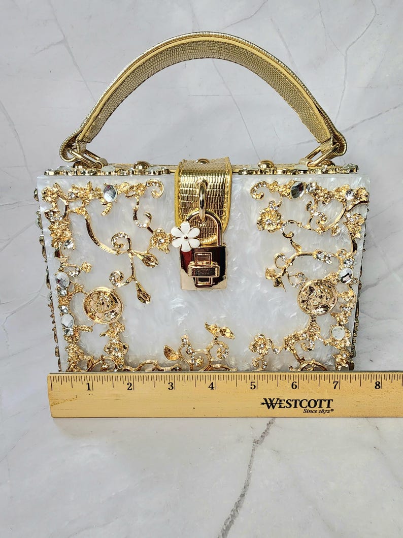 May include: A white box clutch with a gold handle and ornate gold and crystal embellishments. The clutch has a gold lock and a small white flower charm. The clutch is 6 inches wide.