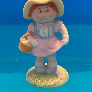 May include: A ceramic figurine of a child with red hair wearing a yellow sun hat, a pink dress with blue trim, and white shoes. The child holds a small woven basket in one hand and stands on a textured, circular base painted to resemble grass.