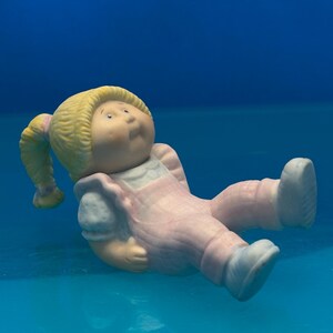 May include: A small figurine of a child with blonde hair styled in two side ponytails, wearing a light pink outfit with white sleeves. The figure has a round face, rosy cheeks, and is depicted in a seated position with legs extended forward.