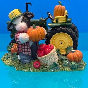 May include: A decorative figurine featuring a cow wearing a John Deere hat, holding a pumpkin, and standing near a green tractor. The scene includes pumpkins, a basket of red apples, and a grassy base. The figurine has a blue background.