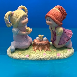 May include: A ceramic figurine featuring two children with round faces kneeling on a grassy base. One child has blonde pigtails and a pink outfit, holding a yellow chick. The other has reddish-brown hair and a maroon dress, looking at a nest on a tree stump.