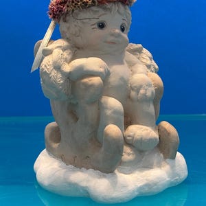 May include: A ceramic cherub figurine, seated on a cloud base. The cherub has a serene expression, adorned with a floral crown. The figurine is painted in soft, neutral tones with subtle details, and has small wings. A decorative piece.
