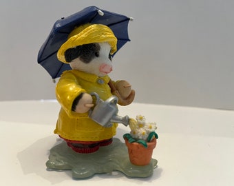 May include: A figurine of a cow wearing a yellow raincoat and hat, holding a blue umbrella. The cow is watering a pot of white daisies with a silver watering can. The figurine is on a green base.