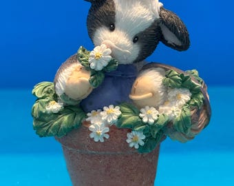 May include: A small figurine of a black and white calf wearing a blue garment, sitting inside a terracotta-colored flower pot. The calf is surrounded by green leaves and small white daisy-like flowers, holding a white flower near its nose.