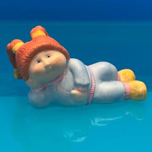 May include: A small figurine of a child with orange hair styled in two pigtails with yellow ties. The figure is lying on its side, wearing a light blue long-sleeved outfit with pink trim at the collar, cuffs, and ankles, and yellow shoes.
