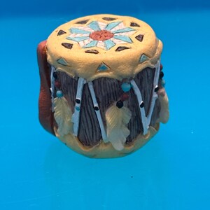 May include: A miniature decorative drum with a yellow top featuring a turquoise and orange floral design. The drum's sides are brown with vertical gray lines, adorned with white strings, feathers, and turquoise beads. The drum is set against a blue background.