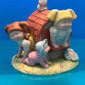 May include: A ceramic figurine featuring three children and a small cat around a brown doghouse. One child with blonde pigtails sits on the roof, another with red pigtails crawls on the side, and a baby in a blue bonnet crawls on the grassy base.