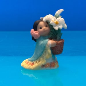 May include: A small figurine of a girl holding a pot of white flowers. The girl wears a blue dress with yellow and orange accents, a pink flower in her hair, and has dark hair. The figurine is set against a blue background.