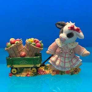 May include: A figurine of a cow wearing a pink dress and a red bow, pulling a green wagon filled with apples. The wagon has two baskets of apples. The figurine is on a bed of autumn leaves.