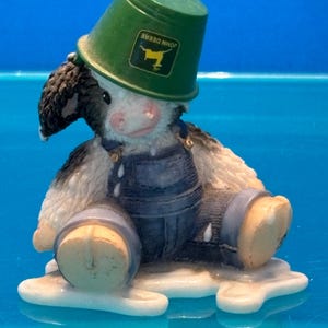 May include: A figurine of a cow wearing blue overalls and a green bucket hat with the John Deere logo. The cow is sitting in a puddle of white liquid, possibly milk. The figurine has black and white markings.