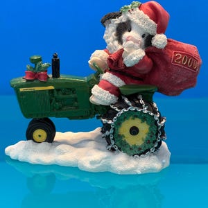 May include: A figurine of a black and white cat dressed as Santa Claus driving a green John Deere tractor. The cat wears a red Santa hat and suit, holding a red sack with "2000" on it. The tractor sits on a snowy base.