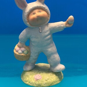 May include: A ceramic figurine of a child wearing a white bunny suit with long ears, holding a small woven basket filled with pastel-colored eggs in one hand and a single egg in the other, standing on a textured green base with a small pink flower.