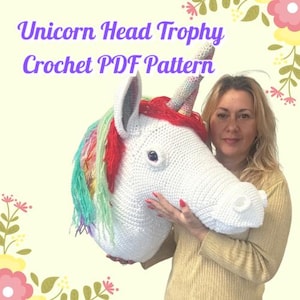 May include: A large, white crochet unicorn head trophy with a rainbow mane and a red accent. The text "Unicorn Head Trophy Crochet PDF Pattern" is displayed in purple. The unicorn is held by a person.