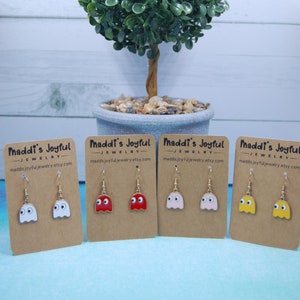 May include: Four pairs of dangle earrings featuring Pac-Man ghosts in white, red, pink, and yellow. The earrings are displayed on brown cards with the text "Maddi's Joyful Jewelry" and a website address.