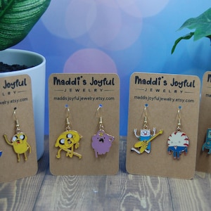 May include: A collection of gold-toned dangle earrings featuring cartoon characters. Each pair is attached to a brown card with the text "Maddi's Joyful Jewelry" and a website address. The earrings showcase various colorful designs, including a blue and white character, a yellow character, and a purple character.
