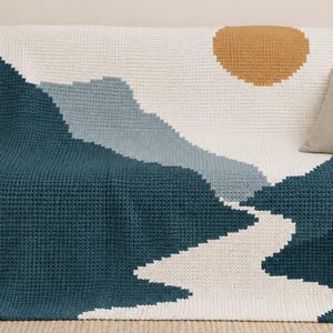 May include: A rectangular crochet blanket featuring a geometric landscape design. The pattern displays a large mustard yellow circle representing a sun against a white background, with layered mountain shapes in dark teal and light blue yarn.