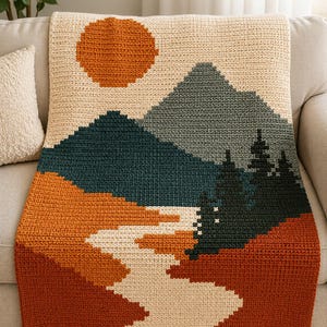 May include: A rectangular crochet blanket featuring a landscape design with a large orange sun, dark teal mountains, a winding cream-colored river, and dark green pine trees against a cream background, all worked in a textured stitch pattern.