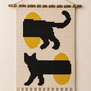 May include: A cream-colored woven wall hanging featuring two black cat silhouettes and two yellow oval shapes. The textile is suspended from a wooden branch with a rope and finished with a row of cream-colored tassels along the bottom edge.
