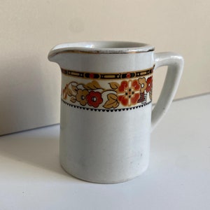 Op de afbeelding: Een kleine, witte keramische creamer met een handvat en tuit. De creamer heeft een decoratieve band van oranje, rode en bruine bloempatronen met zwarte accenten. De rand heeft een gouden rand.
