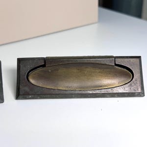 May include: Two antique bronze recessed drawer pulls. Each pull features a rectangular frame with a central oval handle. The metal has a weathered, aged appearance, with variations in color from dark bronze to a lighter brassy tone.